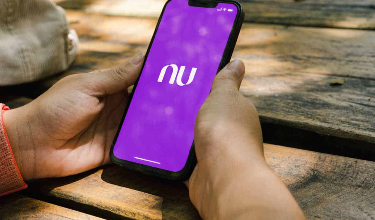 Pessoa ao ar livre, segurando o celular com as duas mãos e, na tela, a logo do Nubank