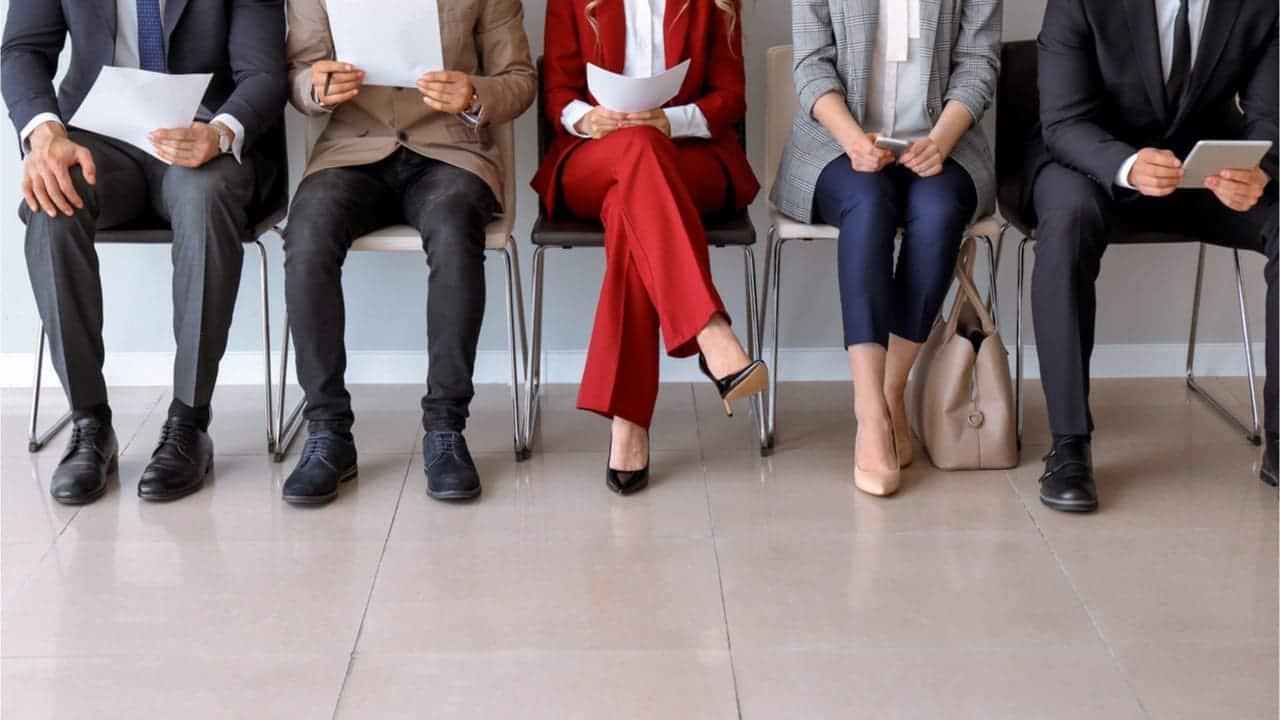 Pessoas sentadas em uma fileira de cadeiras. Elas vestem roupas formais e seguram papéis. Parecem estar esperando algo. A imagem está cortada na altura do busto delas