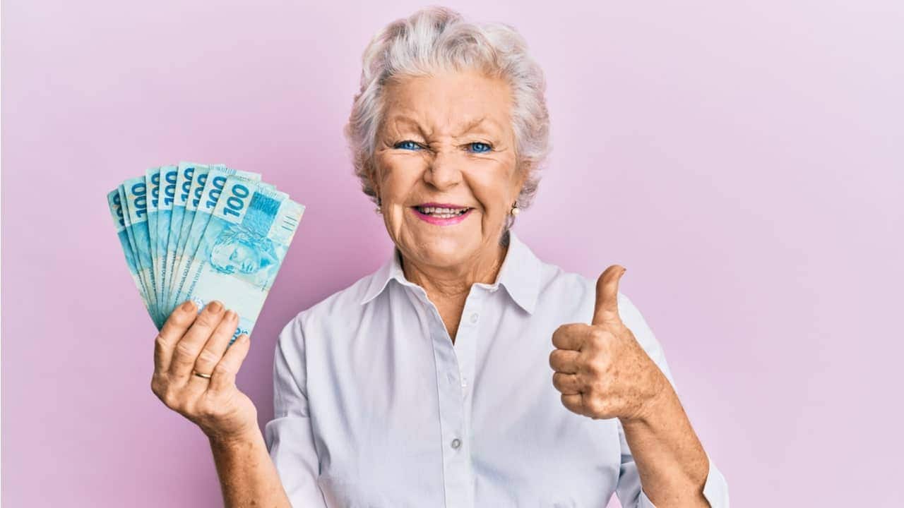 idosa segurando várias notas de R$ 100 em formato de leque com a mão direita. Não mão esquerda, ela faz sinal de "joia"