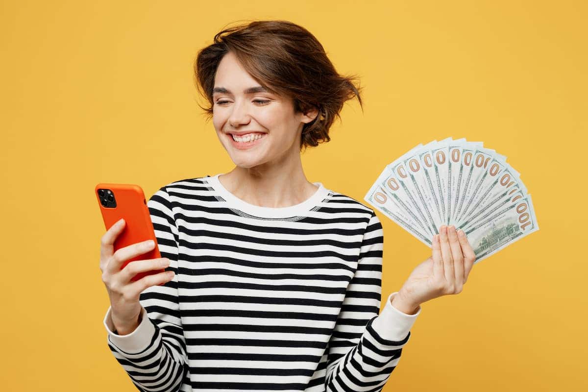 Mulher branca com sorriso no rosto segura leque de dinheiro enquanto usa celular.