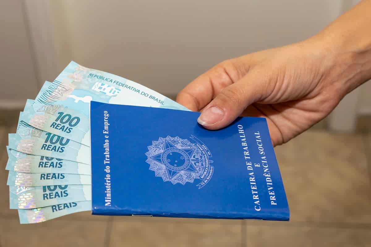 Imagem de uma mĂŁo segurando uma carteira de trabalho com várias notas de 100 dentro, fazendo alusĂŁo ao salário mĂnimo.