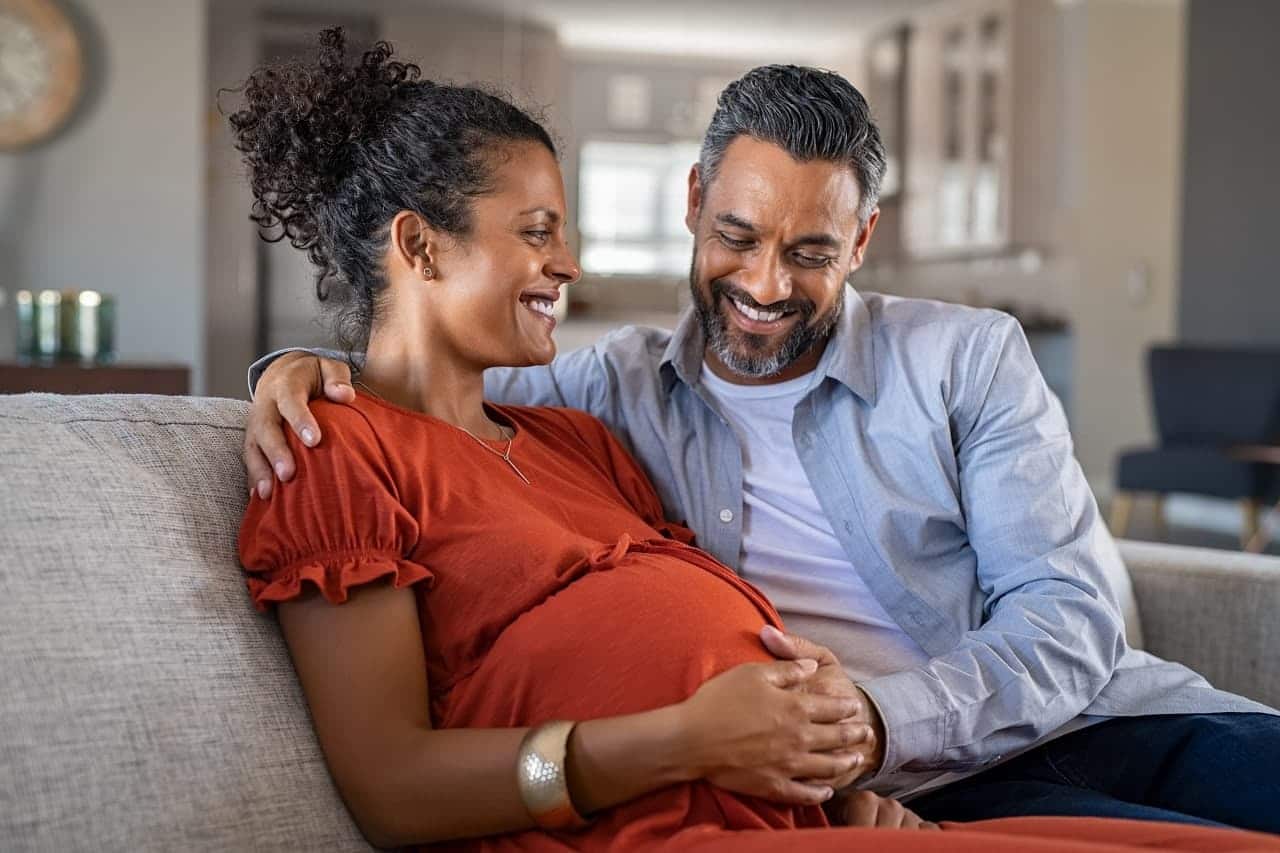 Imagem mostra um casal sorridente sentado no sofá. A mulher está grávida e sorri olhando para o homem, que olha para a barriga com admiração.