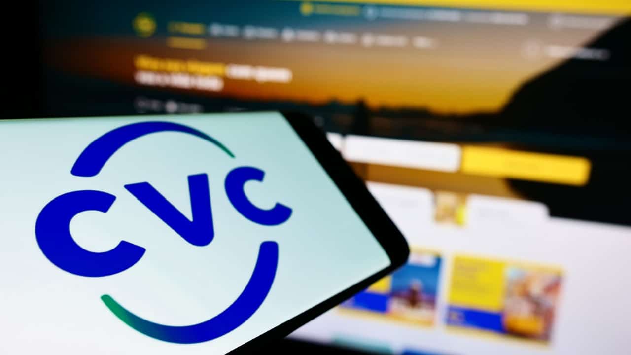 Tela de celular mostrando o logotipo da empresa CVC. Ao fundo, uma tela de computador aparece desfocada, mostrando o site da companhia.