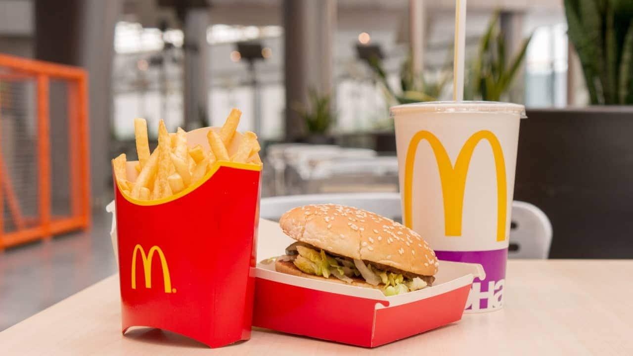 Sobre uma mesa, um combo de refeição do McDonald’s, com sanduíche, batata frita e Coca-Cola. Ao fundo, um ambiente de shopping aparece desfocado.