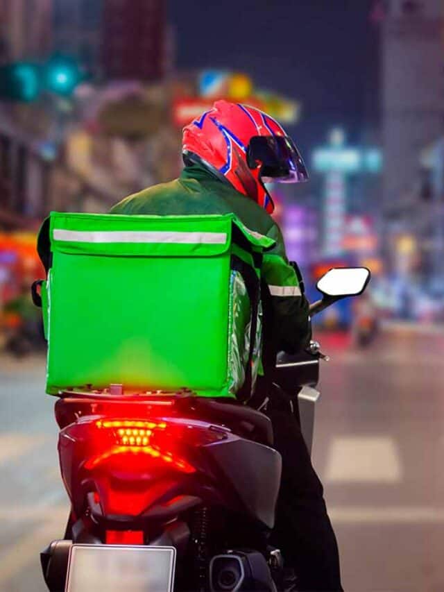 entregador de delivery dirigindo moto numa avenida, ele usa um capacete vermelho e uma bolsa verde na garupa