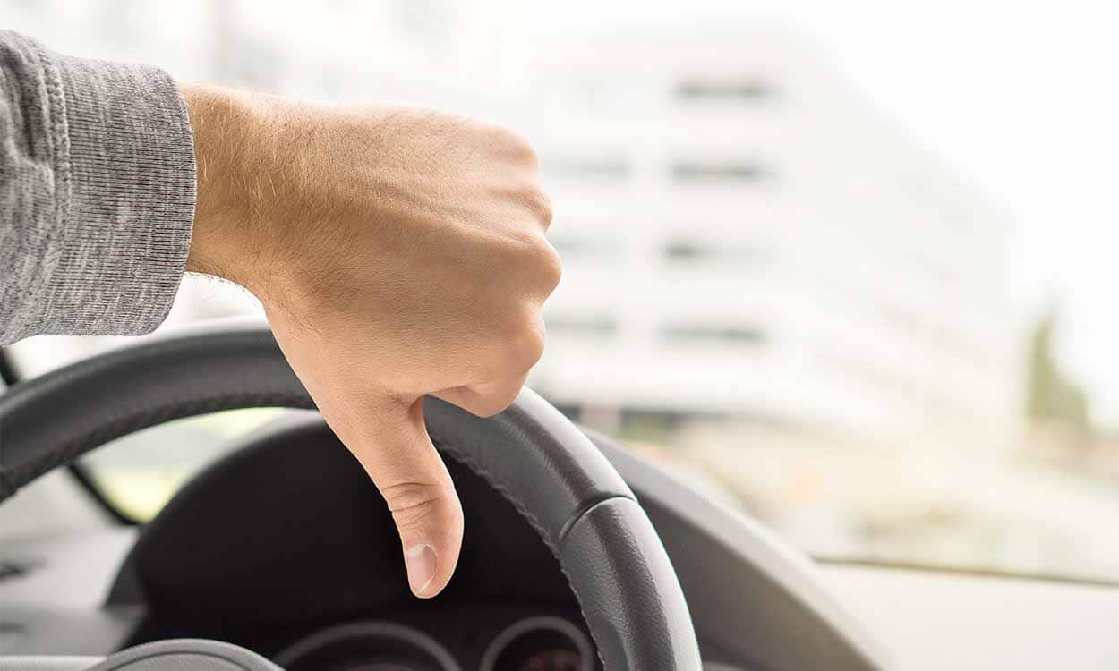 Uma mão com o polegar para baixo, em sinal negativo. Ao fundo o volante indicando que a pessoa está dentro de um carro.
