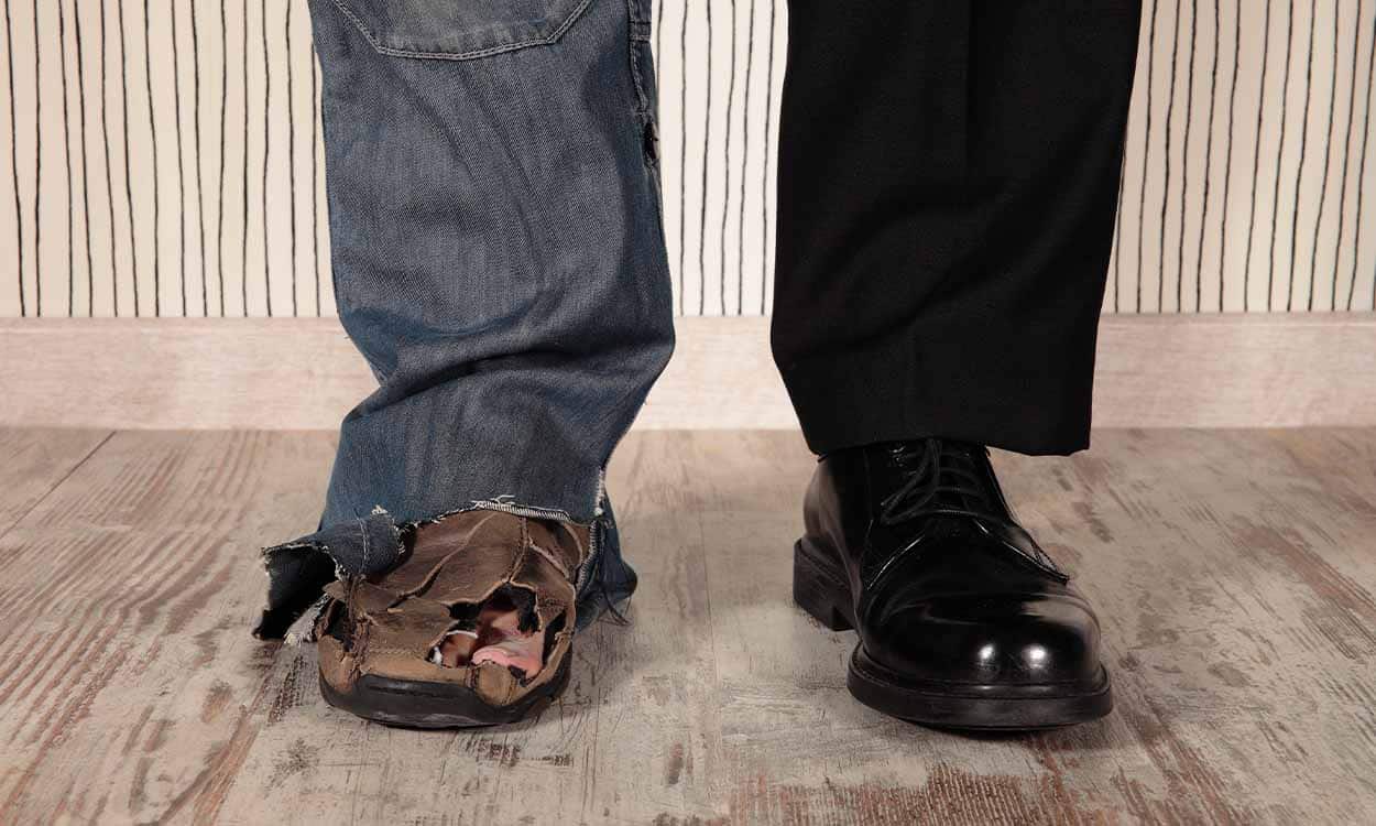 Na imagem, foco em dois pés de uma mesma pessoa. Um com calça jeans e sapato furado, outro com calça e tênis social.