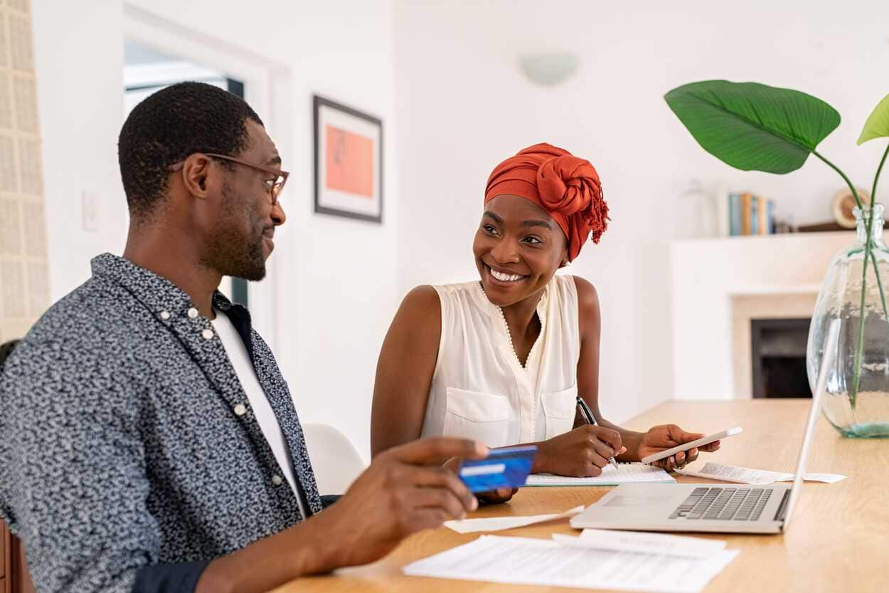 Na imagem um casal está sentado à mesa com um cartão nas mãos e um notebook ligado