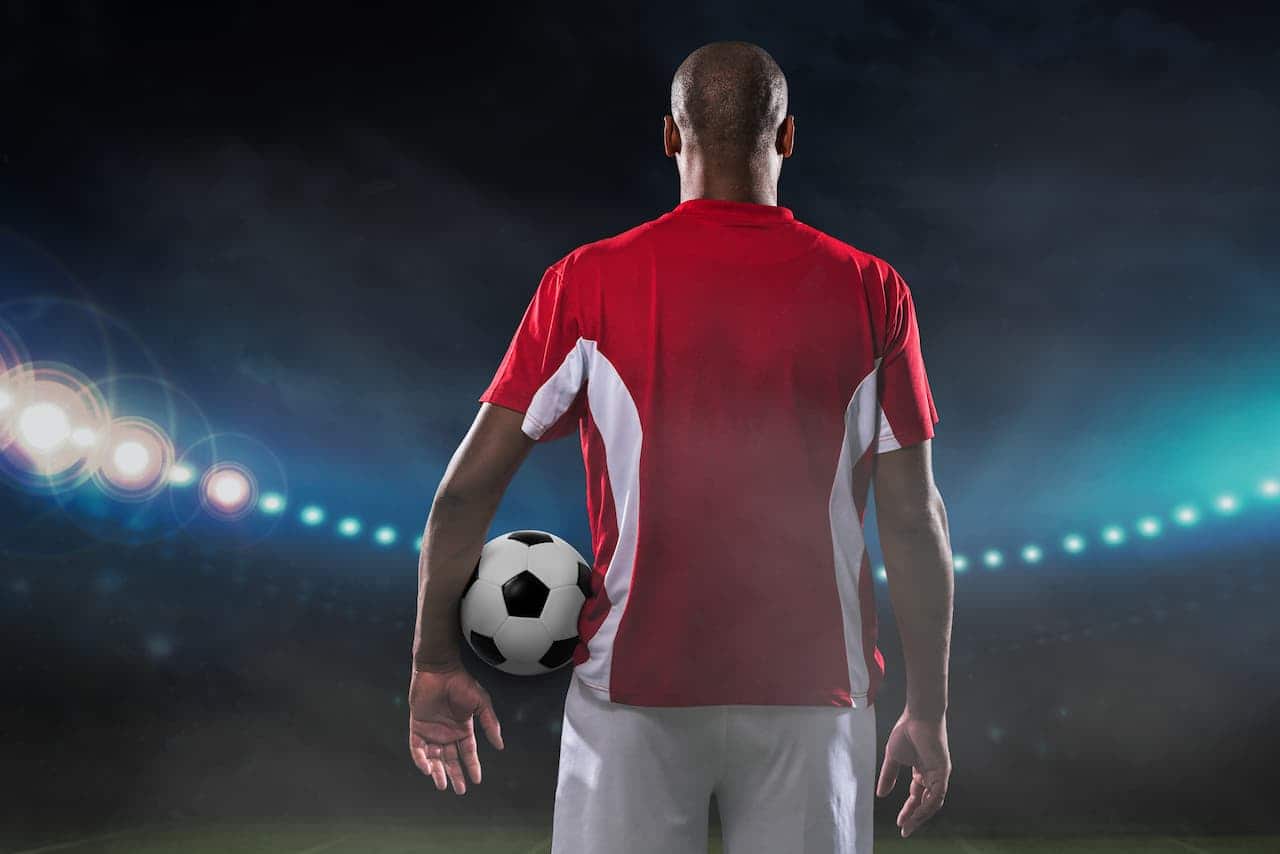 imagem de um jogador de futebol de costas, segurando uma bola, no meio de estádio de futebol