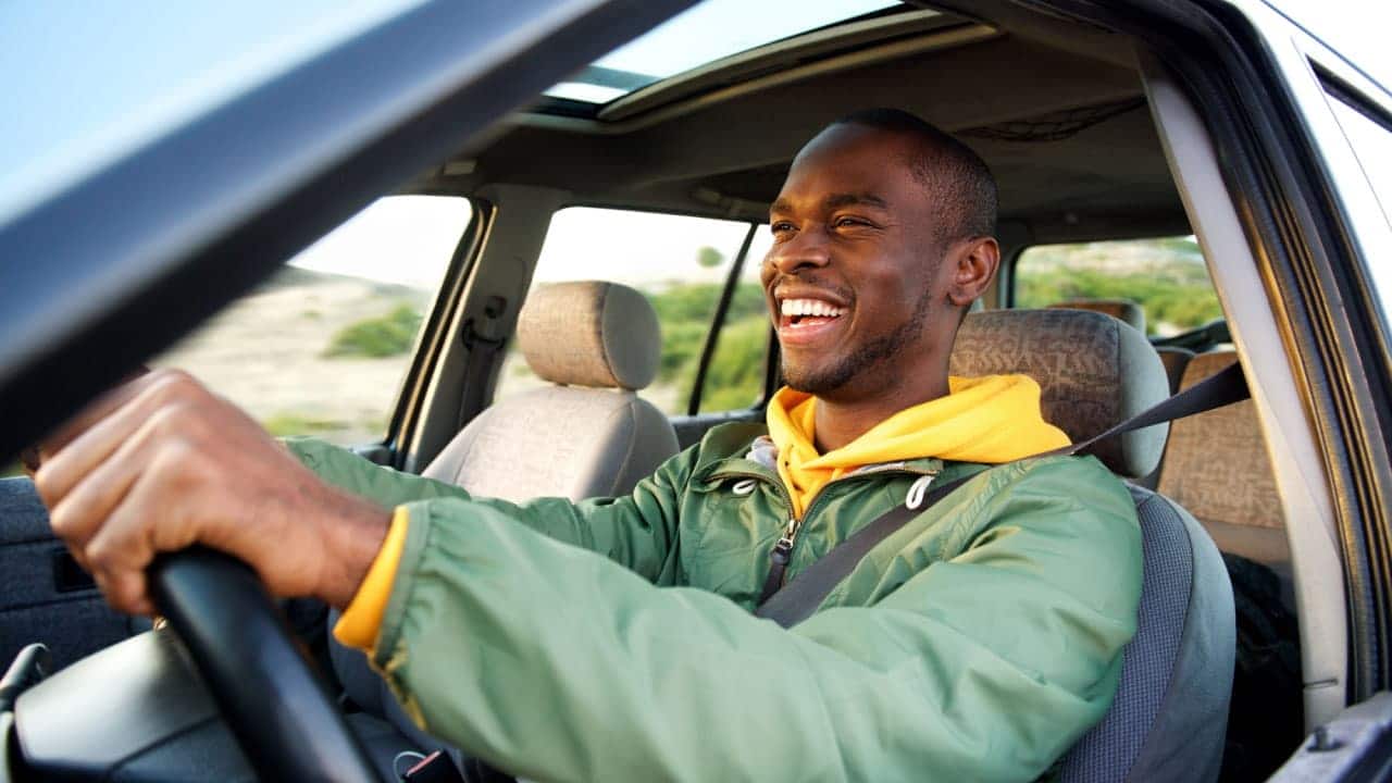 Homem sorridente por ótima notícia para motoristas, dirigindo um carro com o olhar voltado para a estrada à frente.