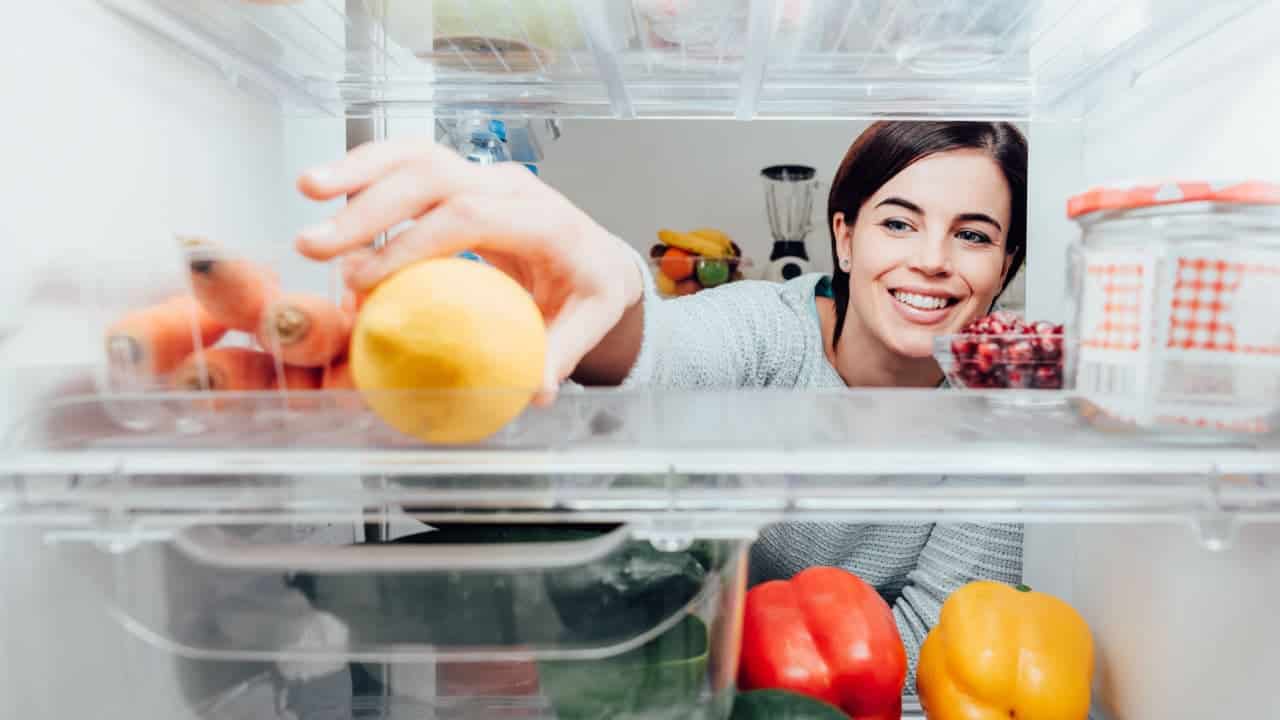 A foto tem a perspectiva de dentro de uma geladeira. Uma mulher está pegando algo que parece uma fruta da geladeira