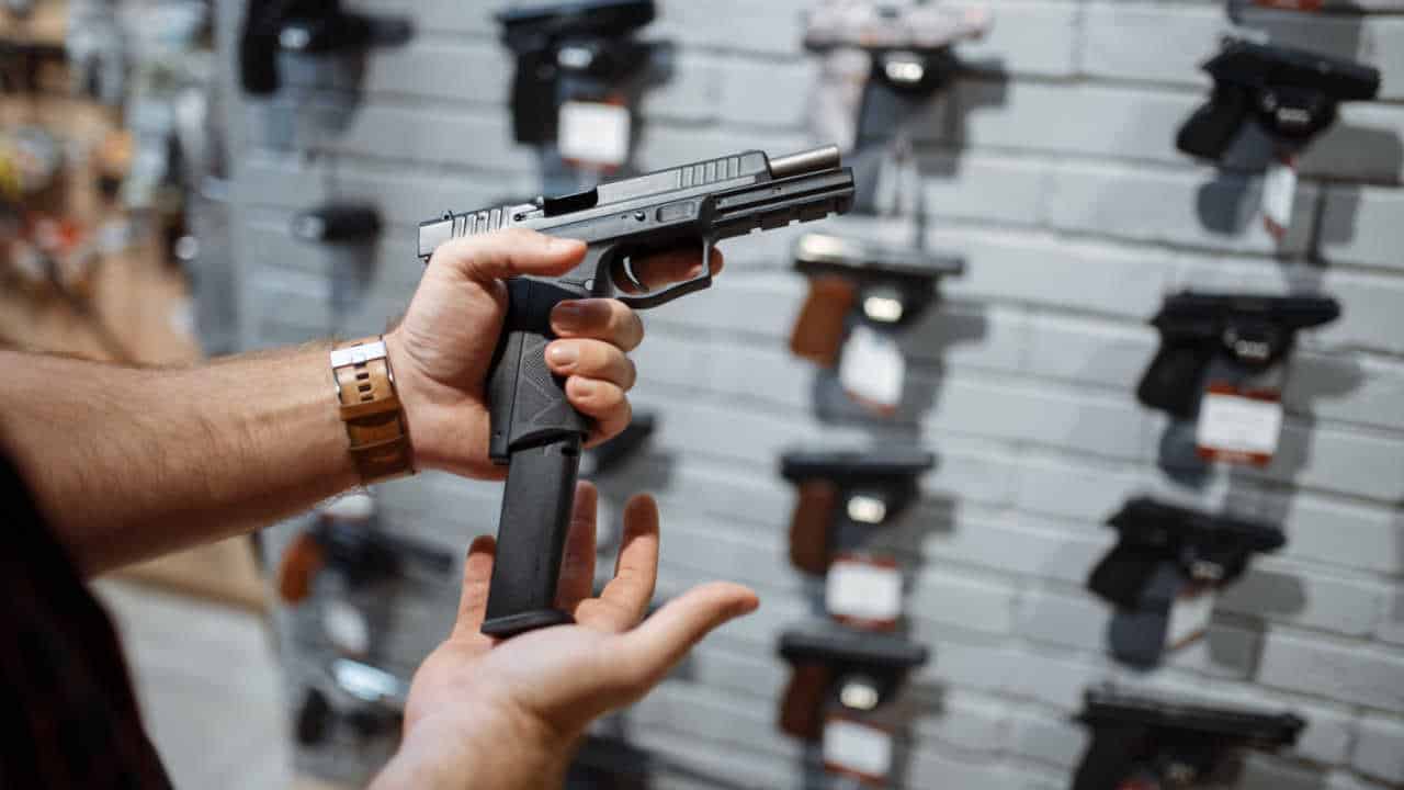Pessoa segurando uma pistola, colocando o cartucho. Ao fundo, há outras armas parecidas em uma parede, penduradas para venda
