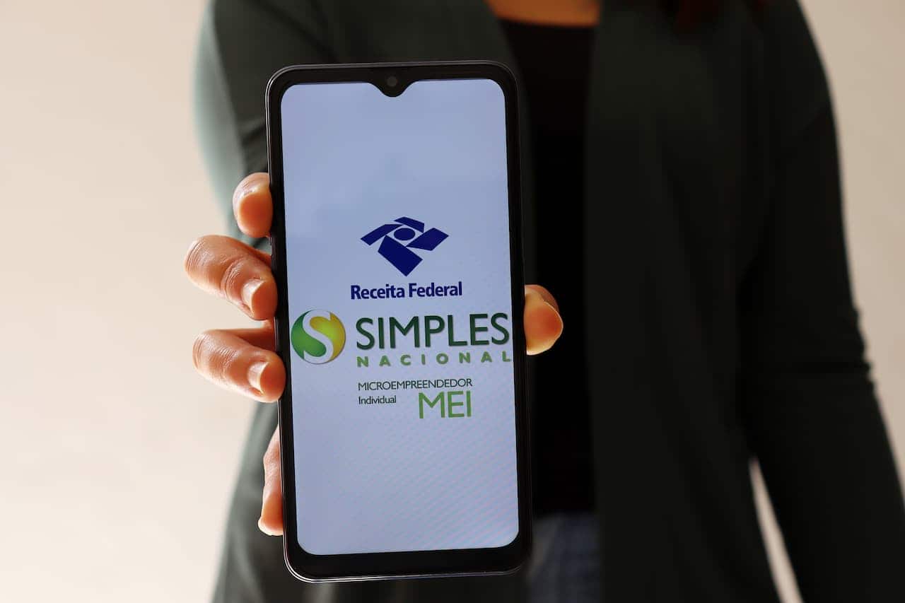 Pessoa segurando um celular apontado para a câmera. Na tela do celular está os logos da Receita Federal, do Simples Nacional e do Microempreendedor Individual (MEI).