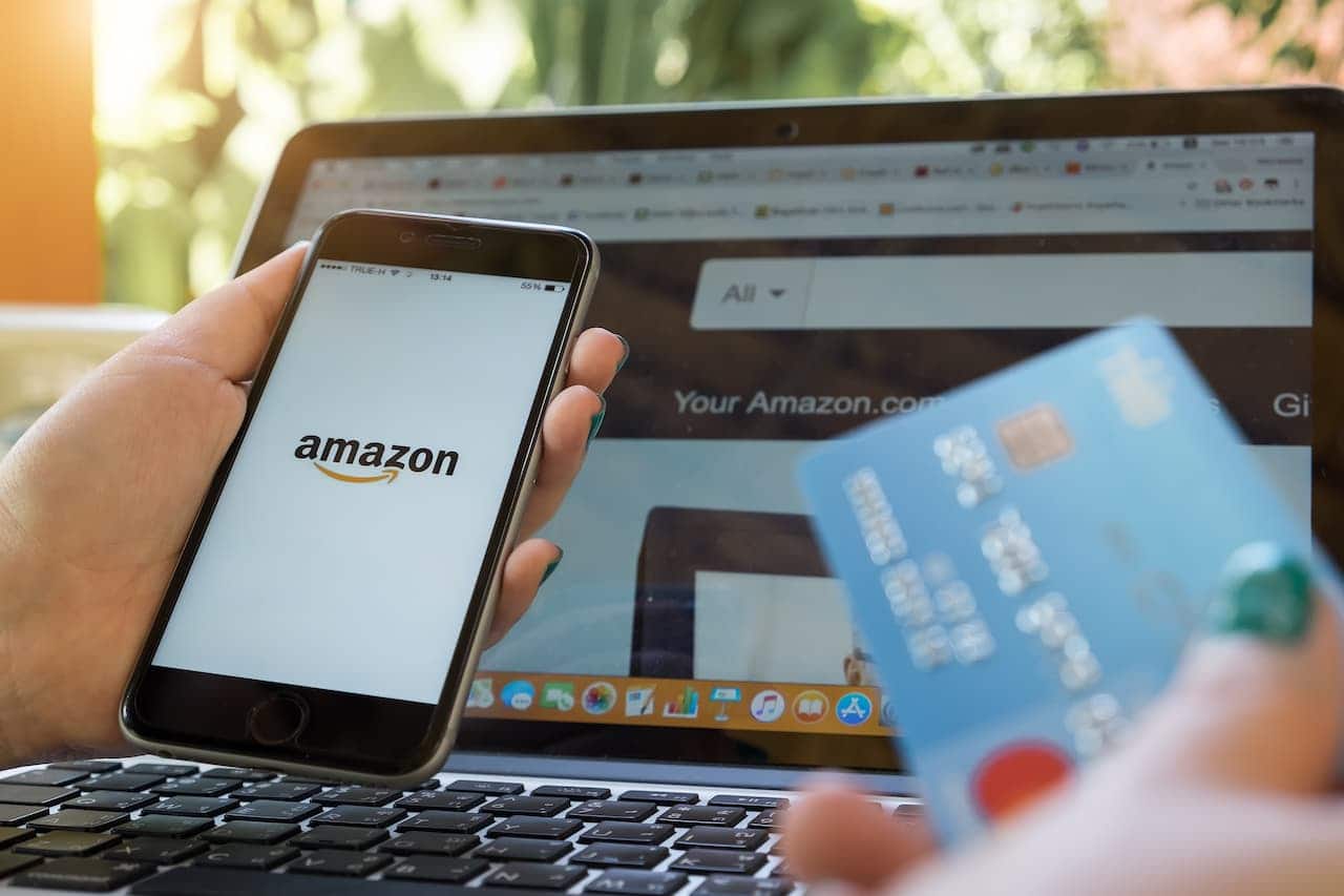 Mãos femininas segurando celular com logo da Amazon na tela, na outra mão um cartão de crédito. Ao fundo um notebook no site da Amazon