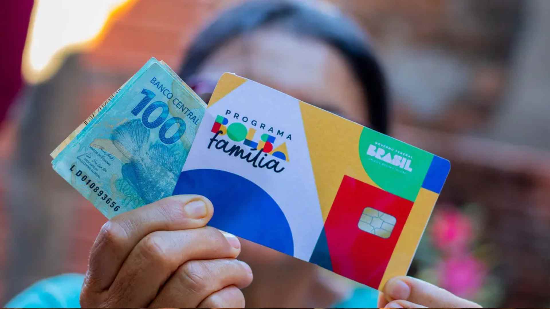 mulher em segurando plano, segurando cartão do bolsa família e uma nota de 100 reais em primeiro plano