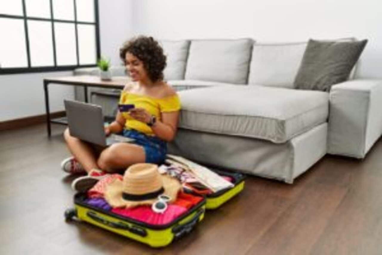 Mulher sentada no chão, com mala de viagem aberta ao lado, segurando cartão de crédito em uma das mãos e com o notebook no colo.