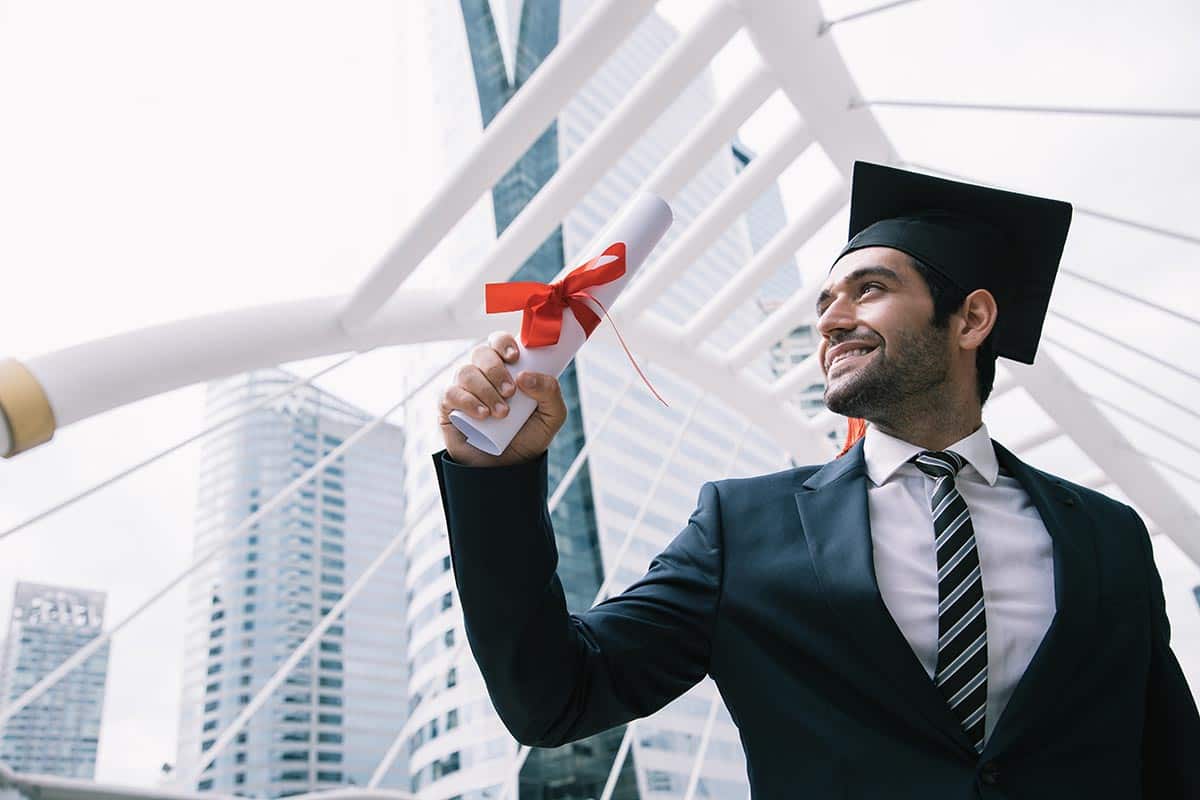 Homem usa capelo de formatura e apresenta diploma em mãos, próximo a uma escada e à paisagem urbana.