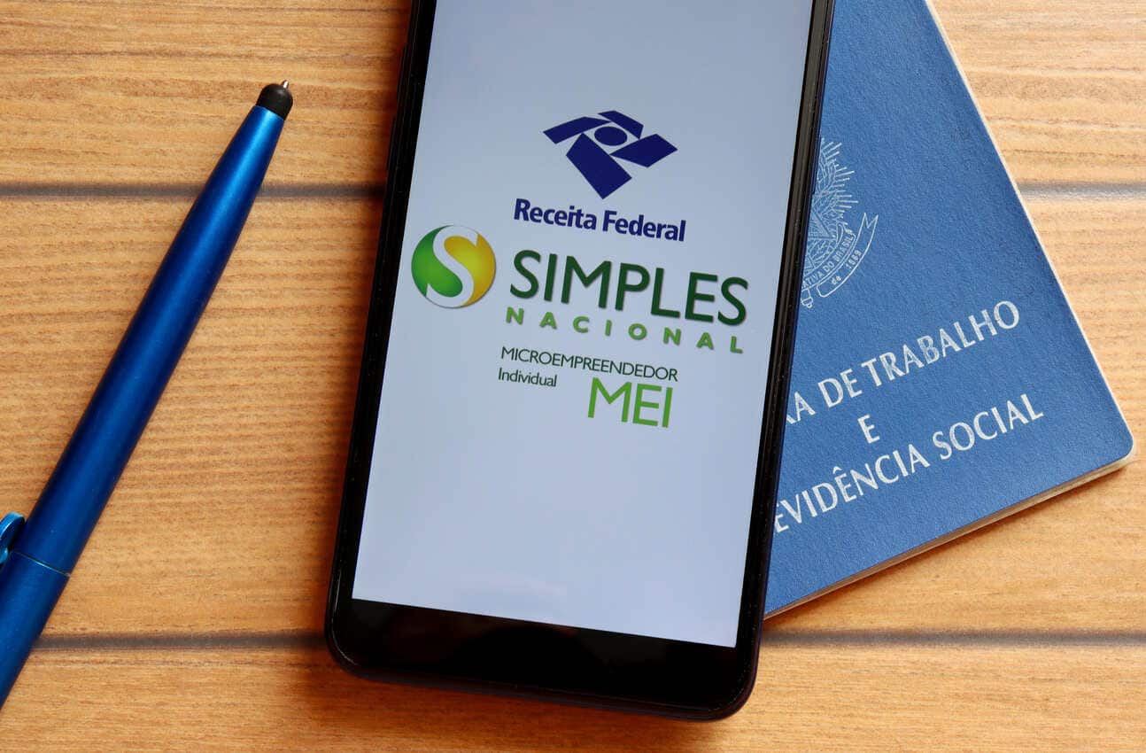 Na imagem, celular com aplicativo do MEI aberto na tela inicial e ao lado uma caneta.