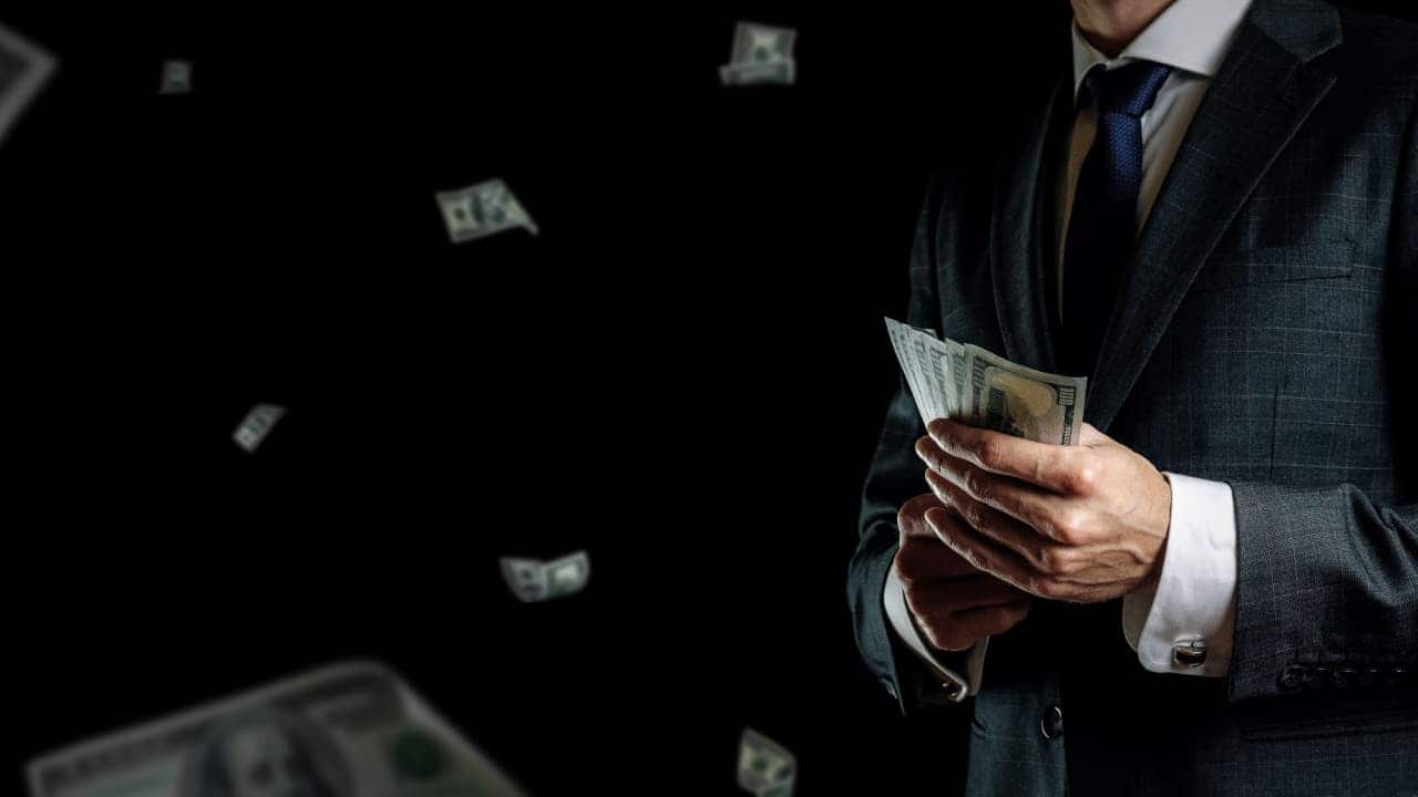 Mão de milionário de terno preto, enquanto segura notas de dólar, ao fundo outras notas também voam pelos ares. ricos