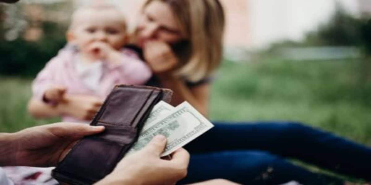 Mãos de uma pessoa segurando carteira e retirando notas de dinheiro. Ao fundo, mãe e bebê aparecem desfocados.