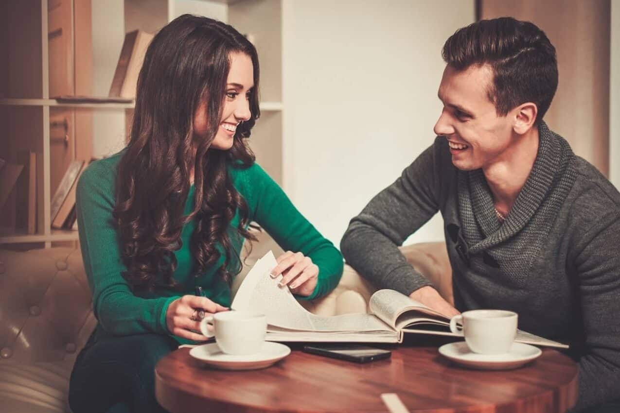 Imagem mostra um casal lendo um livro enquanto tomam café. Eles se olham e sorriem um para o outro.