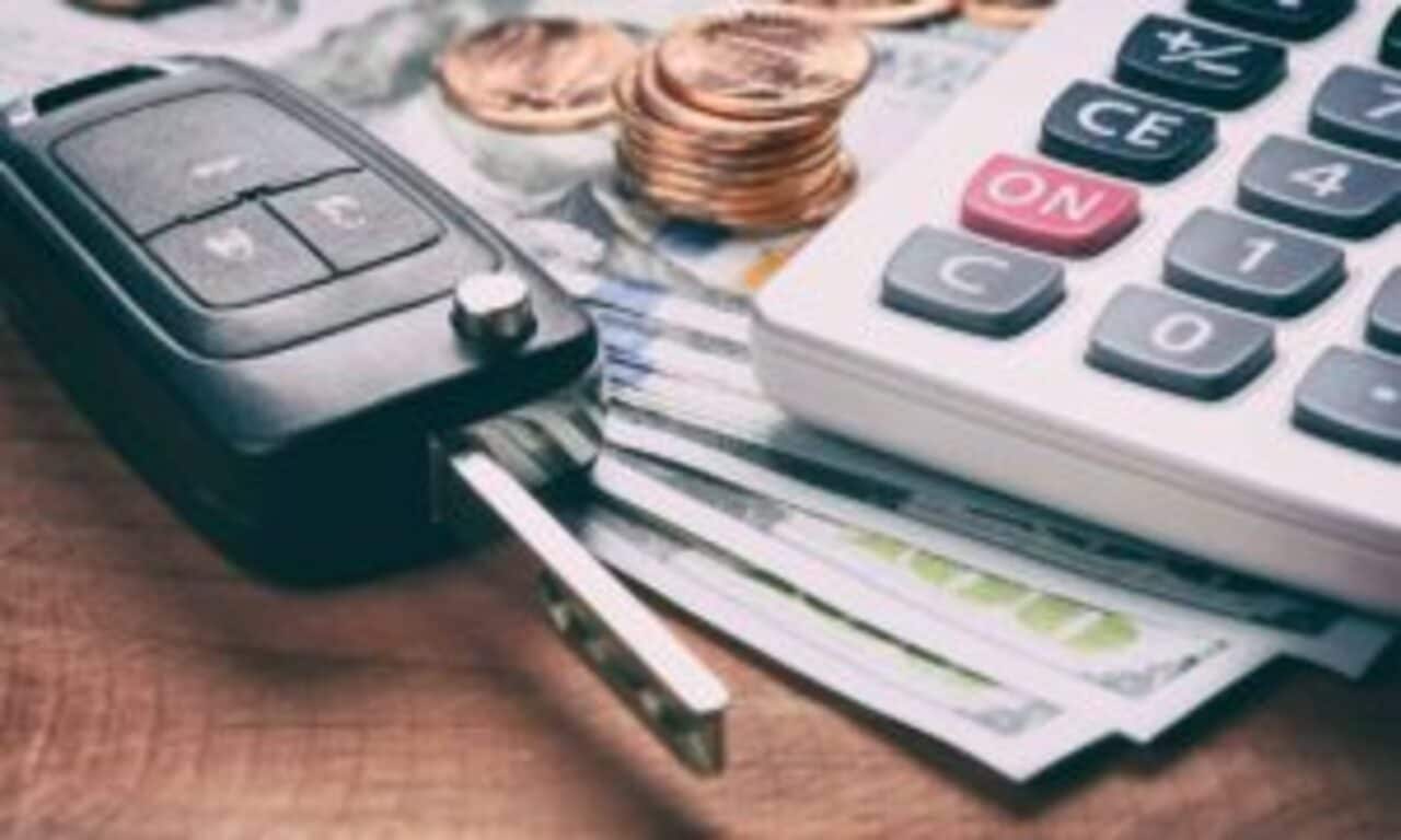 Chave de carro em superfície ao lado de notas de dinheiro, moedas e calculadora.