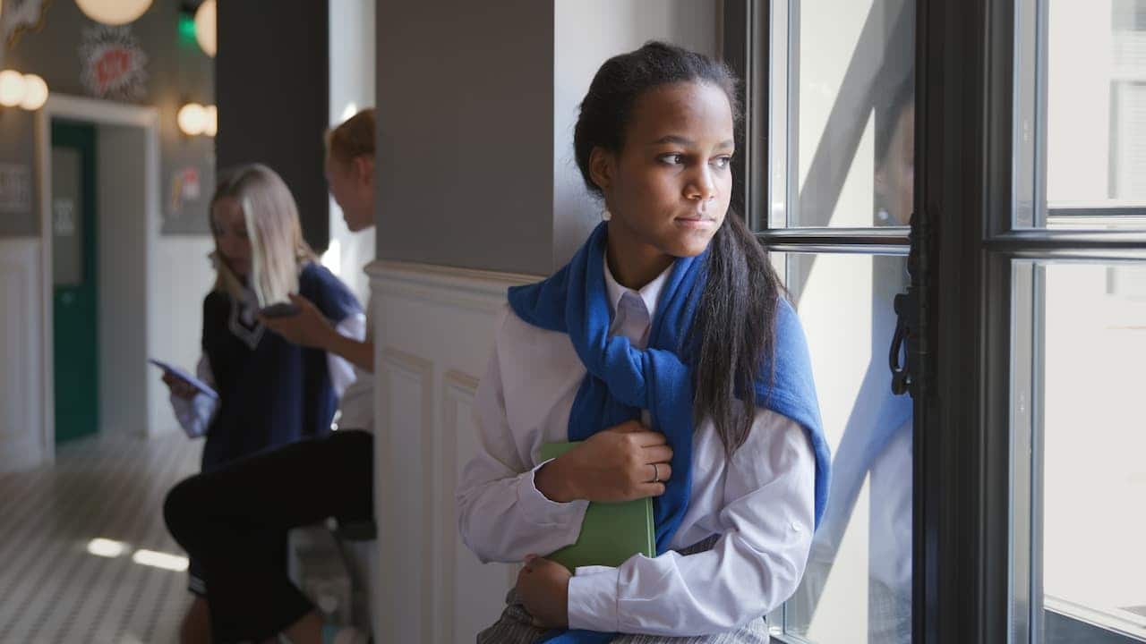 Alunos em um corredor de uma instituição. Uma delas segura um caderno e tem feição preocupada