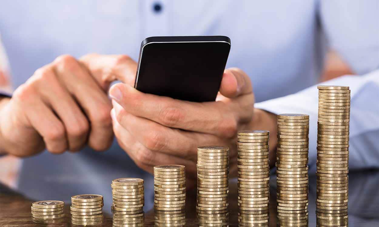 mãos segurando celular em frente a uma pilha de moedas