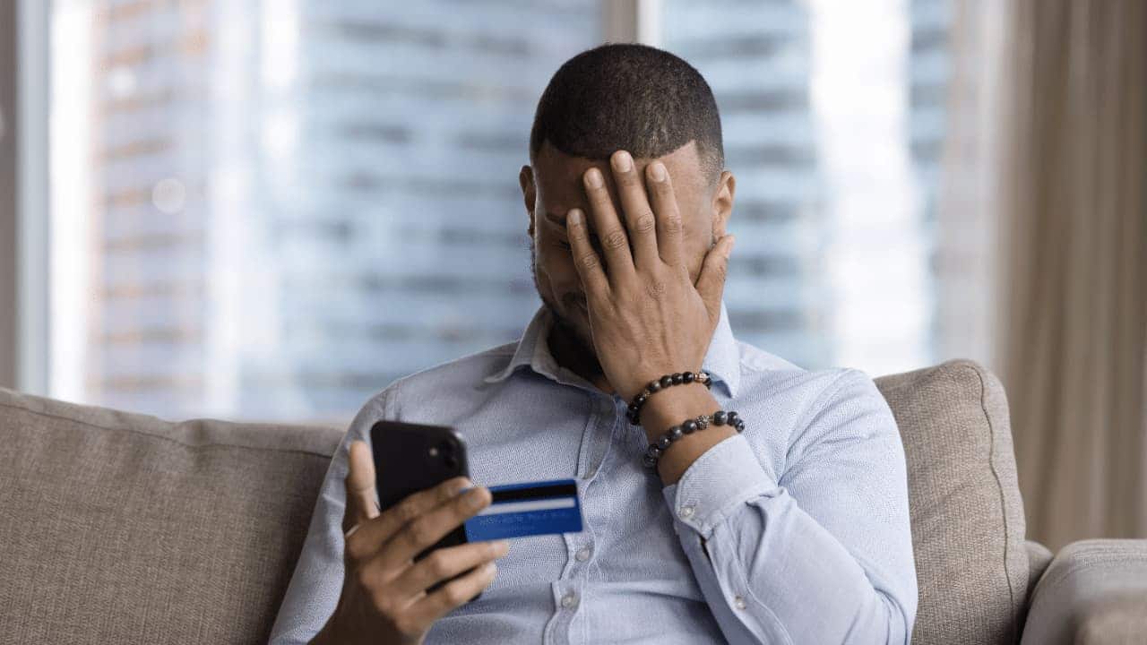 Homem segurando um cartão e um celular, com a mão no rosto decepcionado