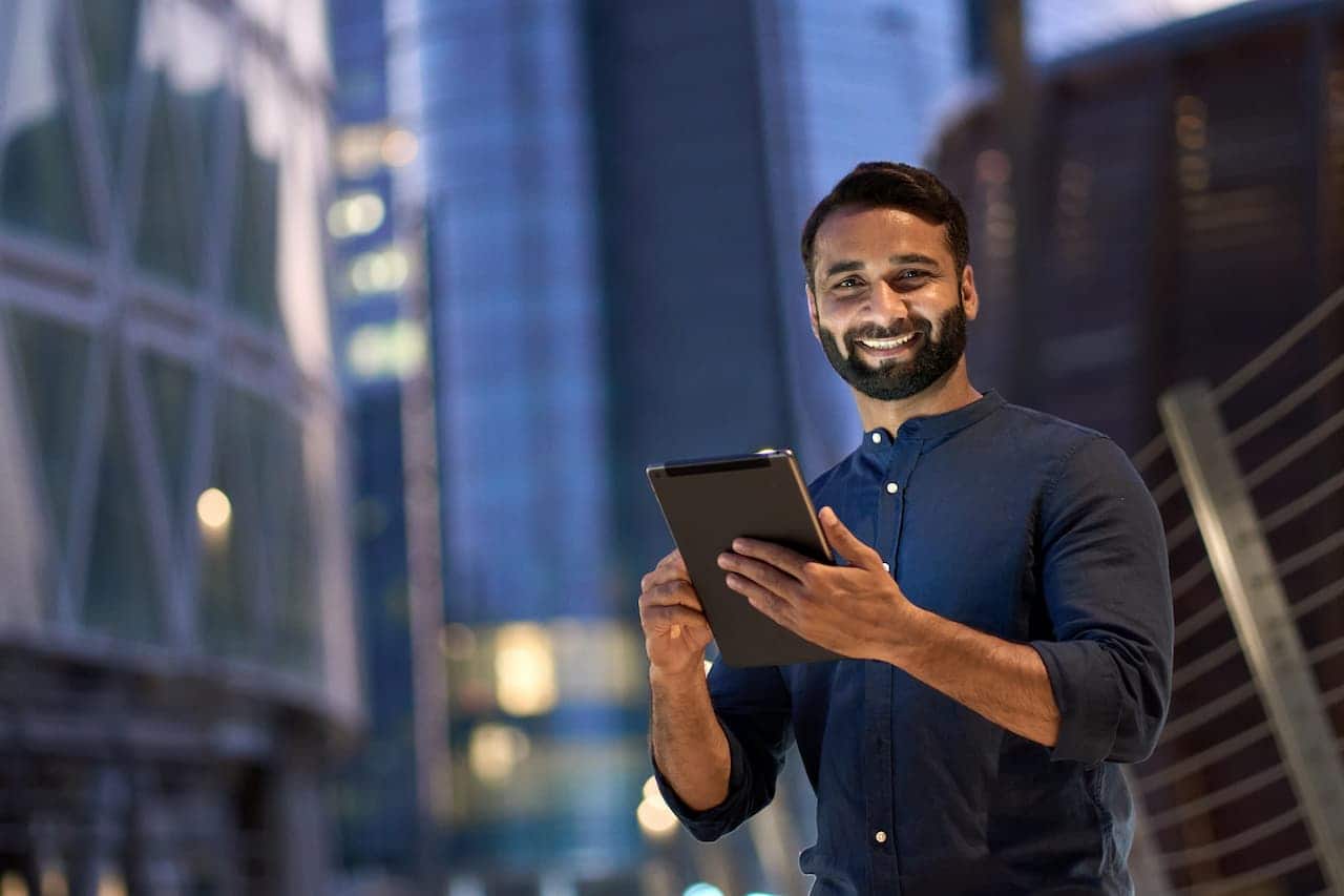 Homem branco com tablet na mão sorrindo. Prédios ao fundo.