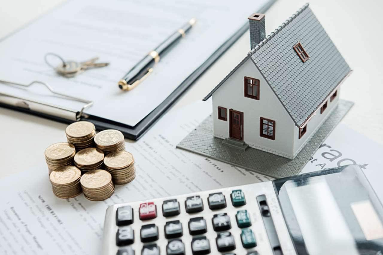 Miniatura de casa rodeada por papéis, uma calculadora, pilhas de moedas, uma caneta e um chaveiro com duas chaves