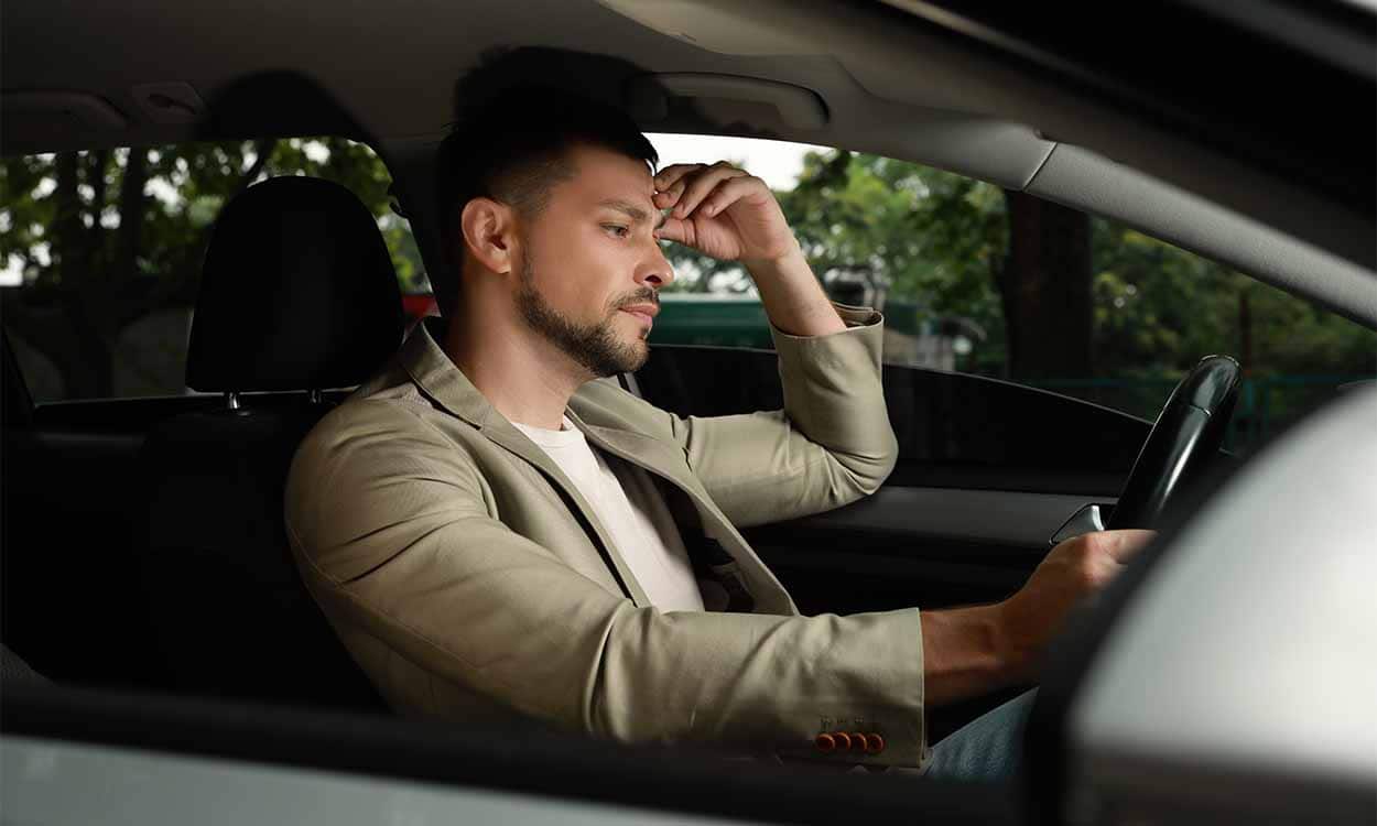 Homem dentro de carro com mão na cabeça em expressão de frustração por ter recebido multas.