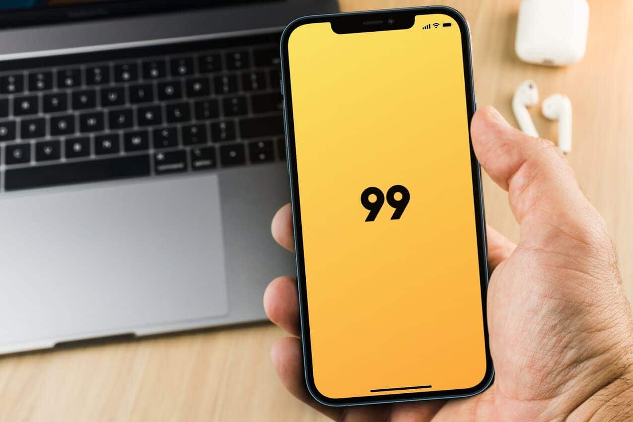 Mão segurando um smartphone que está inicializando o aplicativo do 99. Ao fundo, um notebook aberto