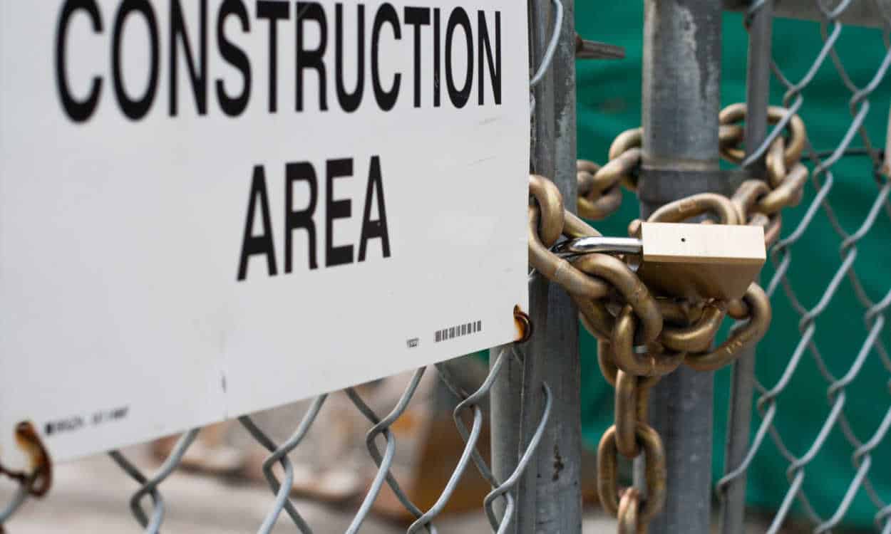 Closeup de portão de um canteiro de obras fechado com uma corrente grossa e uma cadeado. Há uma placa em inglês, com parte ocultada, mas infere-se que está escrito "área de construção"