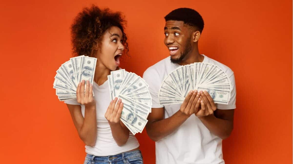 um casal em expressão de choque segura várias notas de dólar, ao fundo tem uma parede vermelha