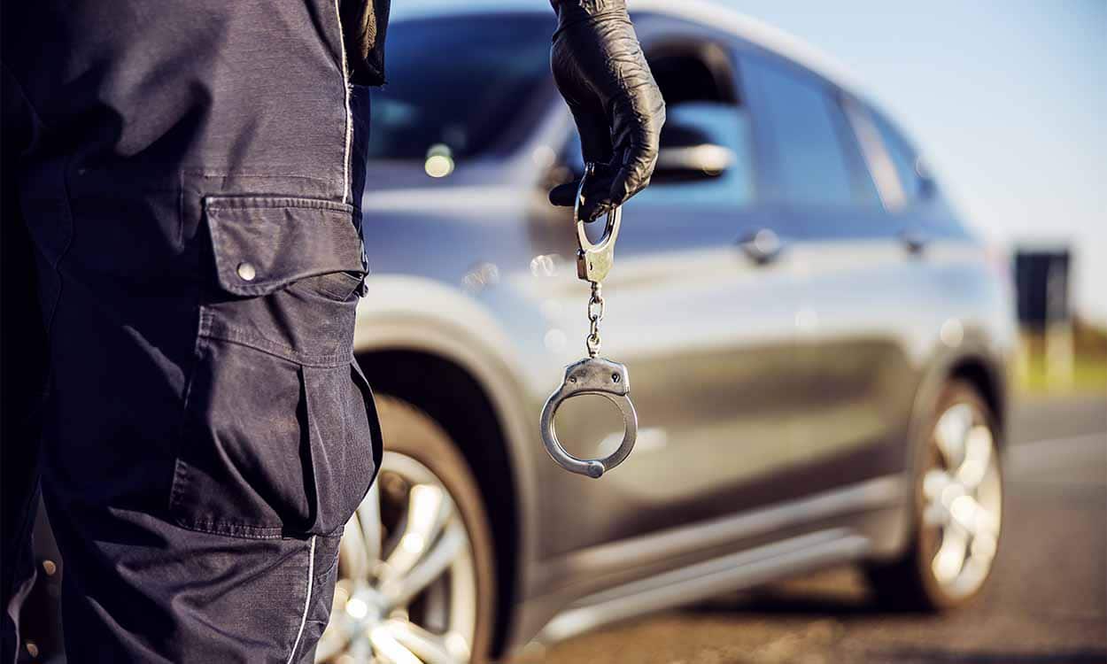 Policial com algema nas mão anda na direção de um carro