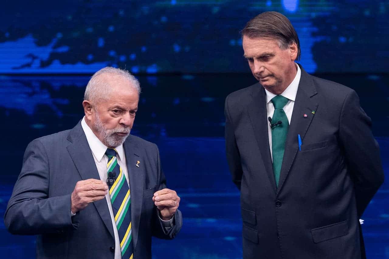 Imagem de Lula e Bolsonaro, lado a lado, debatendo em programa de televisão. Lula gesticula enquanto fala algo, Bolsonaro olha para o chão com as mãos para trás.
