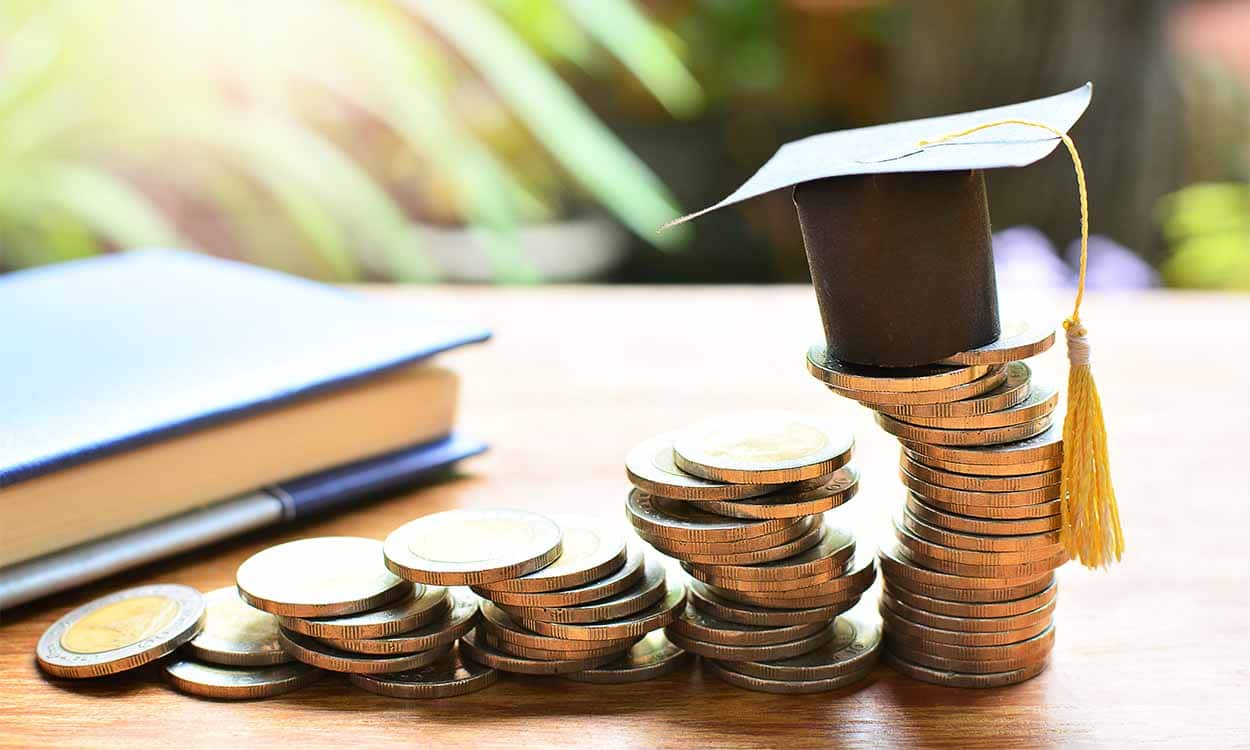 Chapéu de universitário em cima de pilha de moedas, que vai diminuindo em escadinha. Na mesma superfície, há um caderno