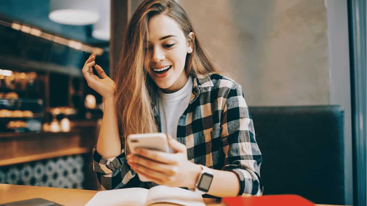 estudante sorri enquanto lê noticia no celular, em cima da mesa tem um livro aberto