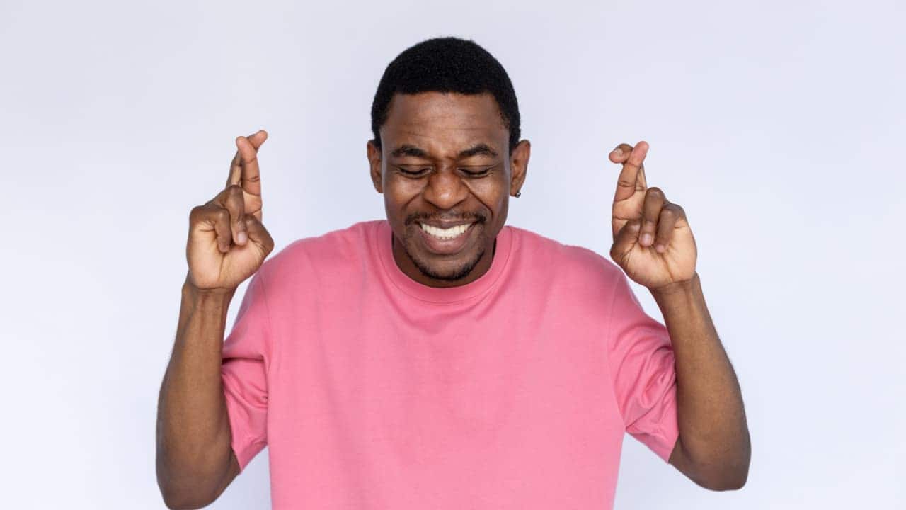 homem feliz com camiseta rosa e olhos fechados enquanto sorri e faz um desejo com os dedos cruzados