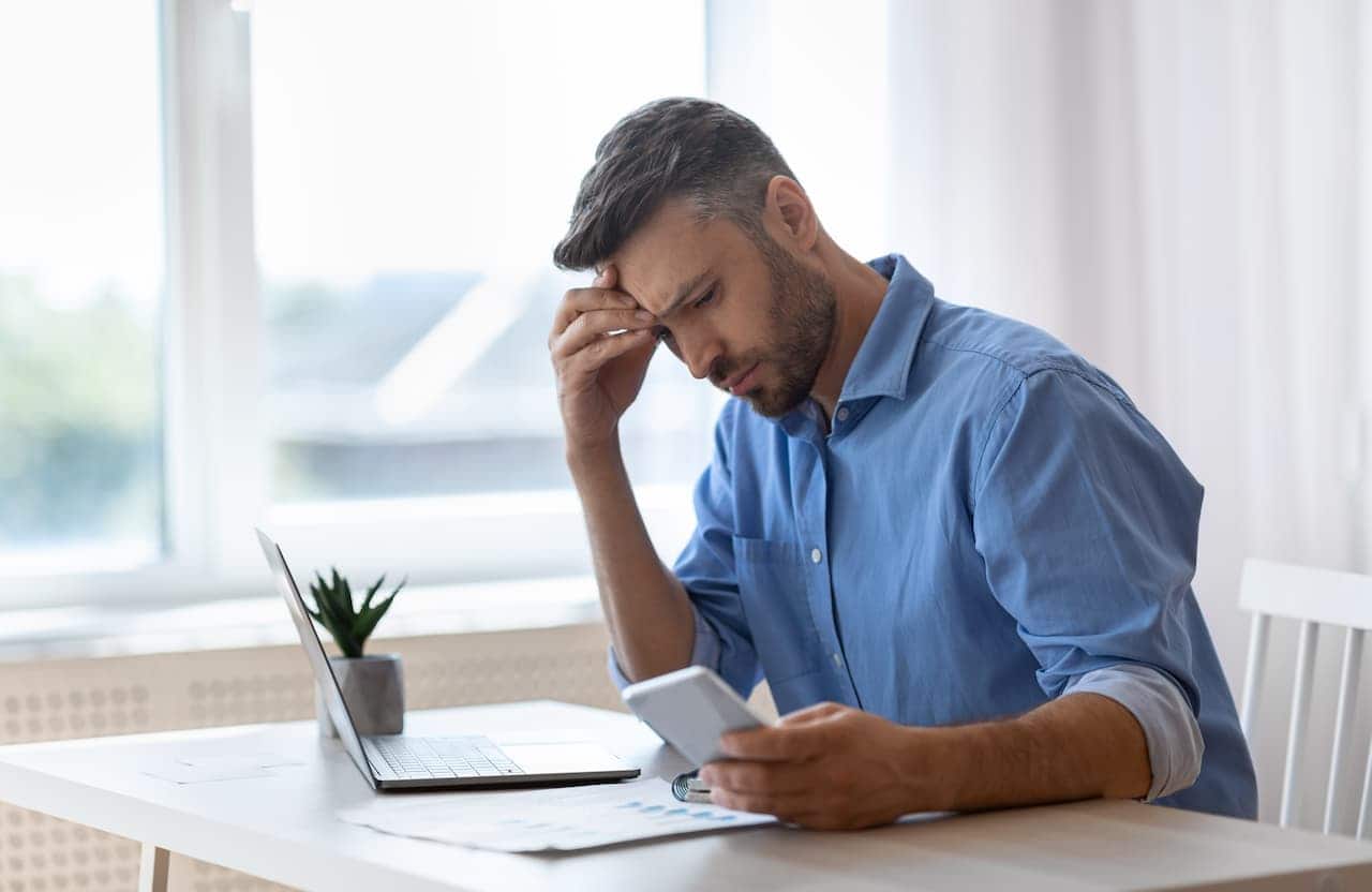 homem preocupado observando celular, com laptop aberto na mesa e uma mão apoiada na testa, em sinal de preocupação