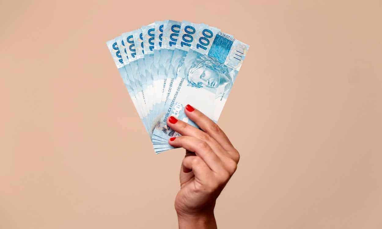 Na imagem, uma mão segurando várias notas de cem reais.