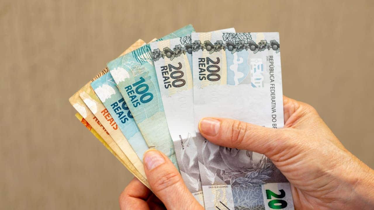 Mão segurando notas de 20, 50, 100 e 200 reais, referetes ao BPC e ao auxílio-inclusão
