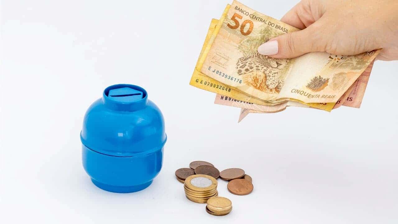 Mão segurando notas de cinquenta reais com miniatura de botijão de gás e moedas ao lado.