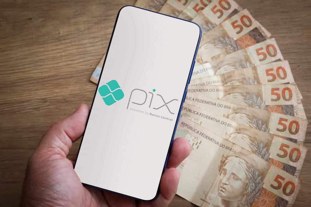 Mão segurando celular com logo Pix. Ao fundo, diversas notas de 50 reais