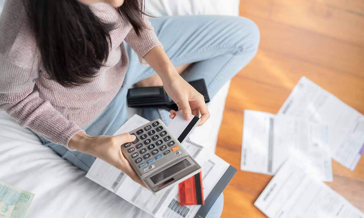 Na imagem, mulher sentada no chão com várias faturas e calculadora.