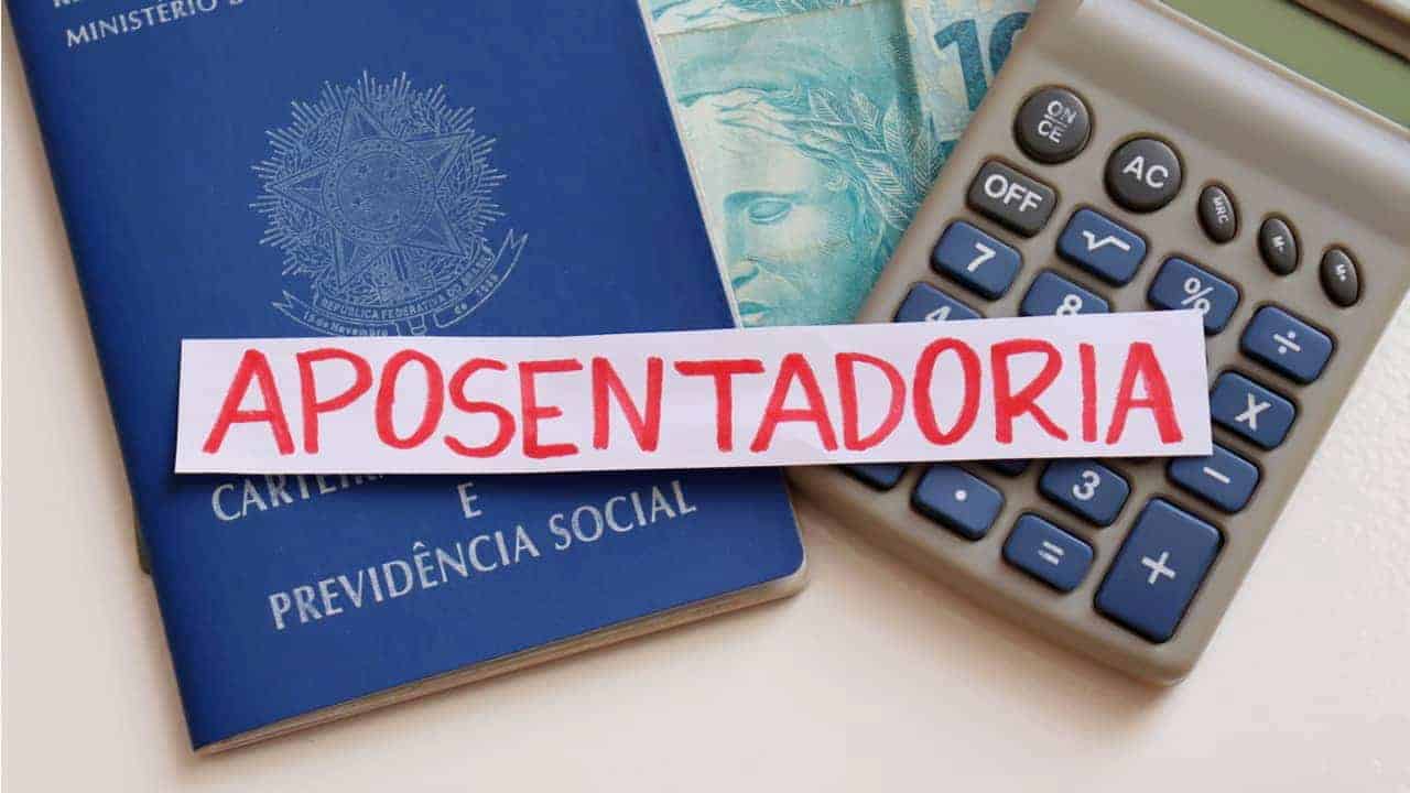 Papel com a palavra "aposentadoria" colocado em cima de uma carteira de trabalho, uma calculadora e uma nota de cem reais.x INSS