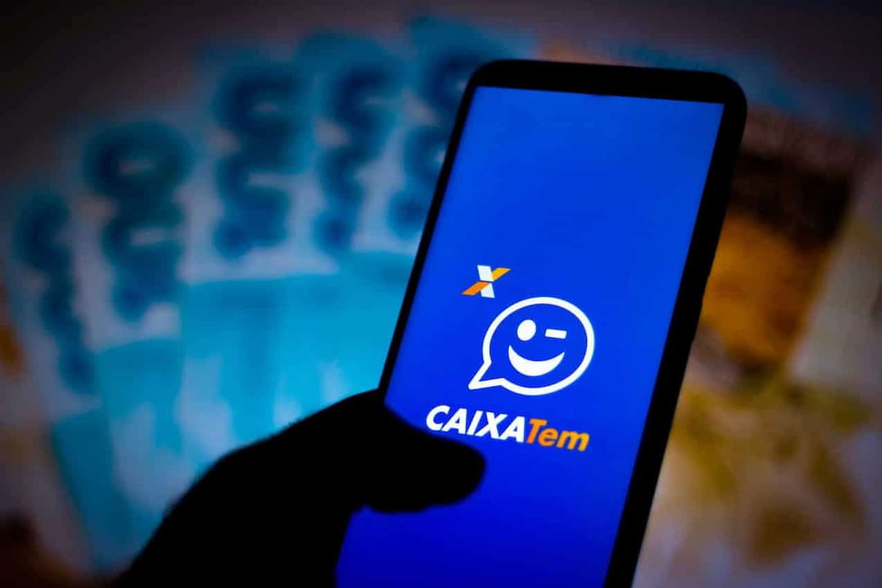 Mão segurando celular com app Caixa Tem aberto. Ao fundo, notas brasileiras embaçadas. Caixa Tem Pix
