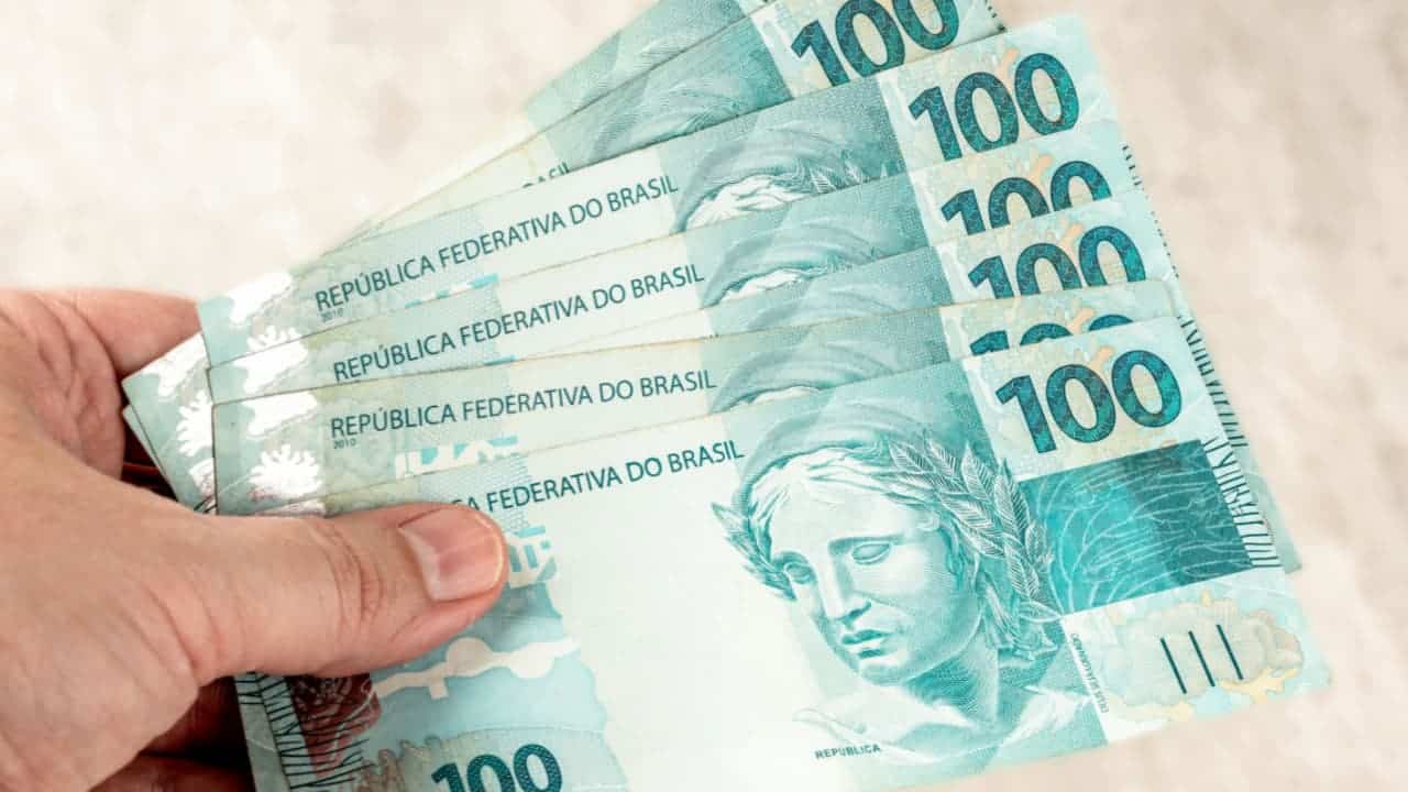 Mão segurando notas de 100 reais.