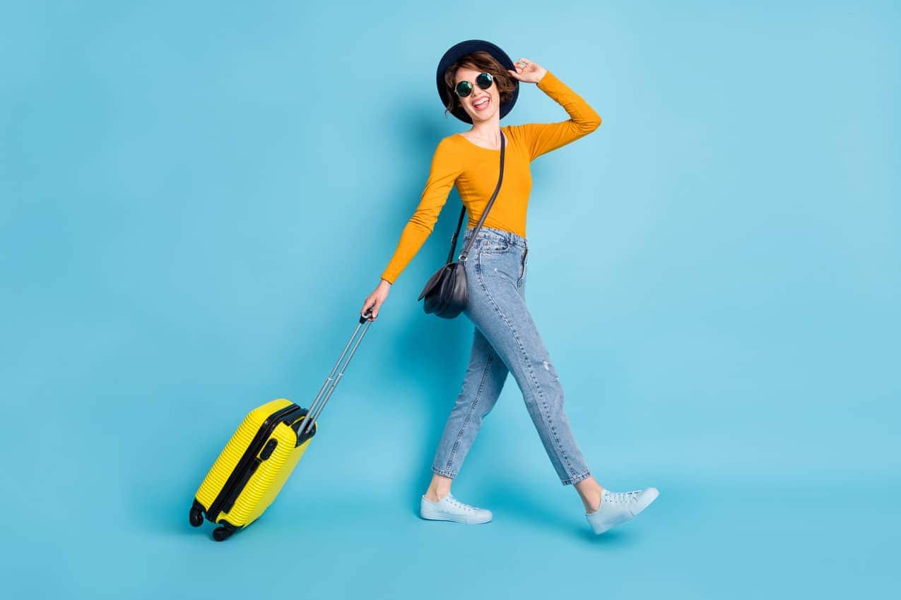 Mulher com chapéu e óculos escuros arrastando uma mala de viagem.